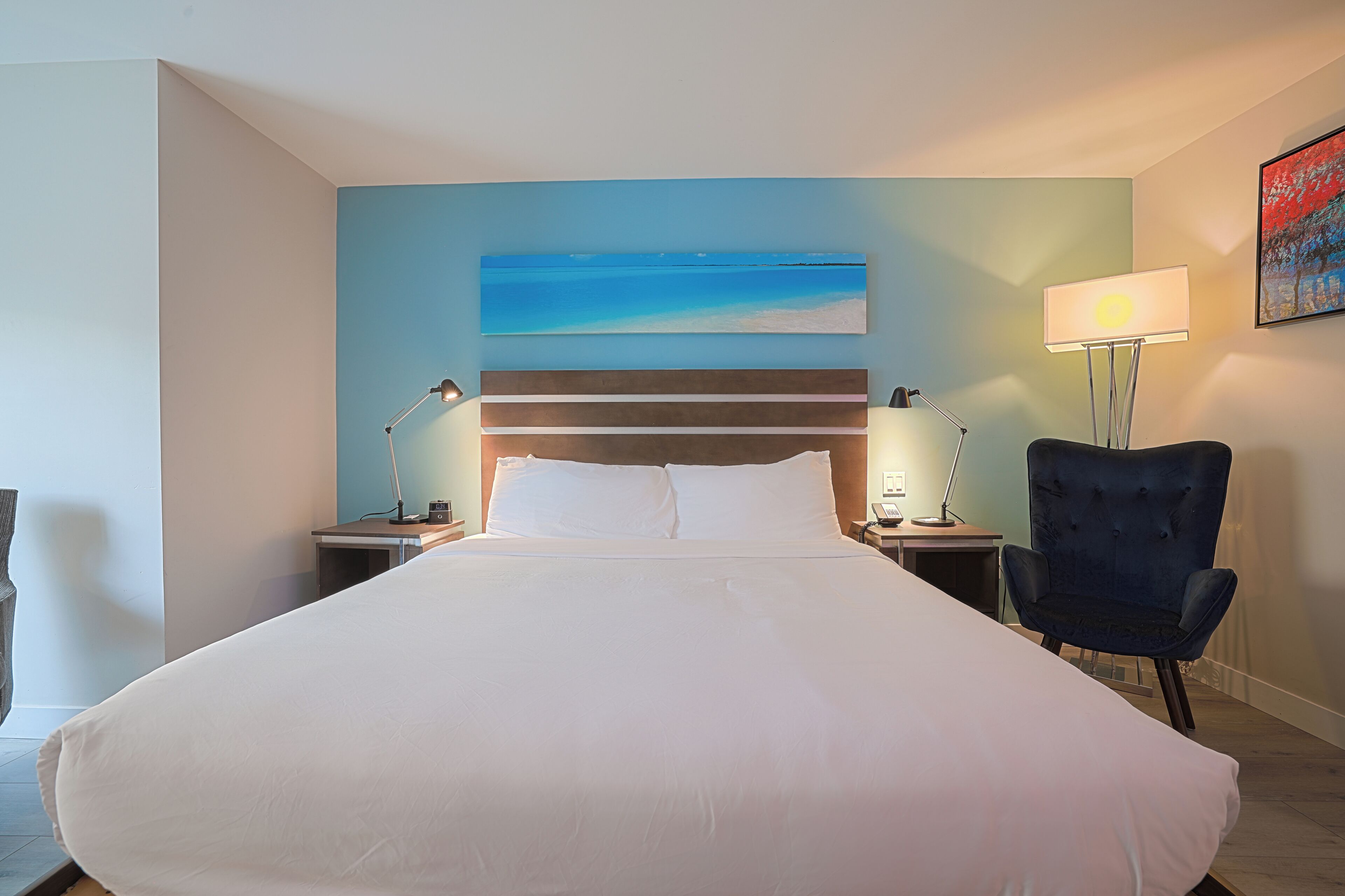 Habitación de hotel con una cama grande, dos mesitas de noche, una silla, un cuadro y una lámpara.