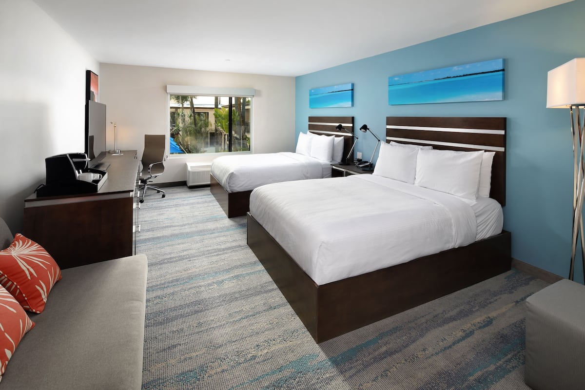 A hotel room with two beds, a desk with a computer, a chair, a lamp, and a view of the outdoors.