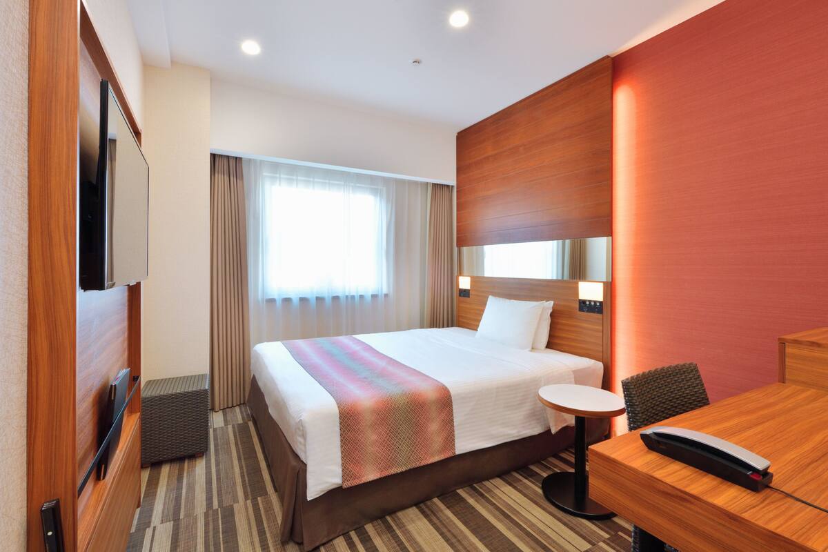 Habitación de hotel con una cama grande, una mesita redonda pequeña, un escritorio, un televisor y una ventana con cortinas.