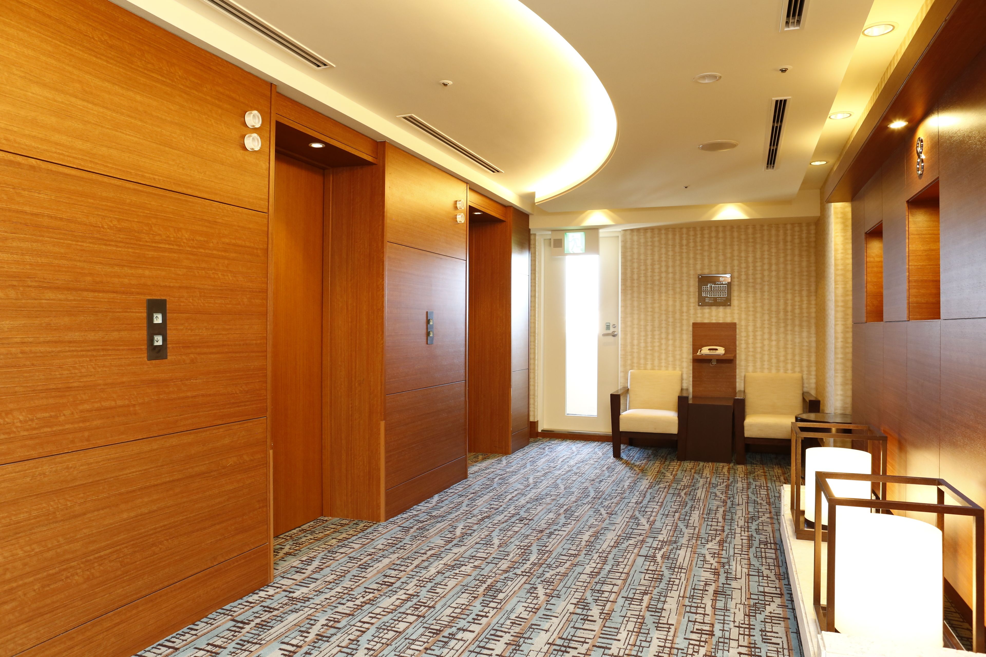 Un vestíbulo de hotel con paneles de madera, alfombra estampada y una zona de estar con sillas y mesas.