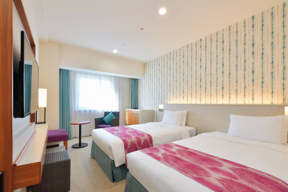 Habitación de hotel con dos camas, un escritorio, una silla, un televisor y una ventana con cortinas.