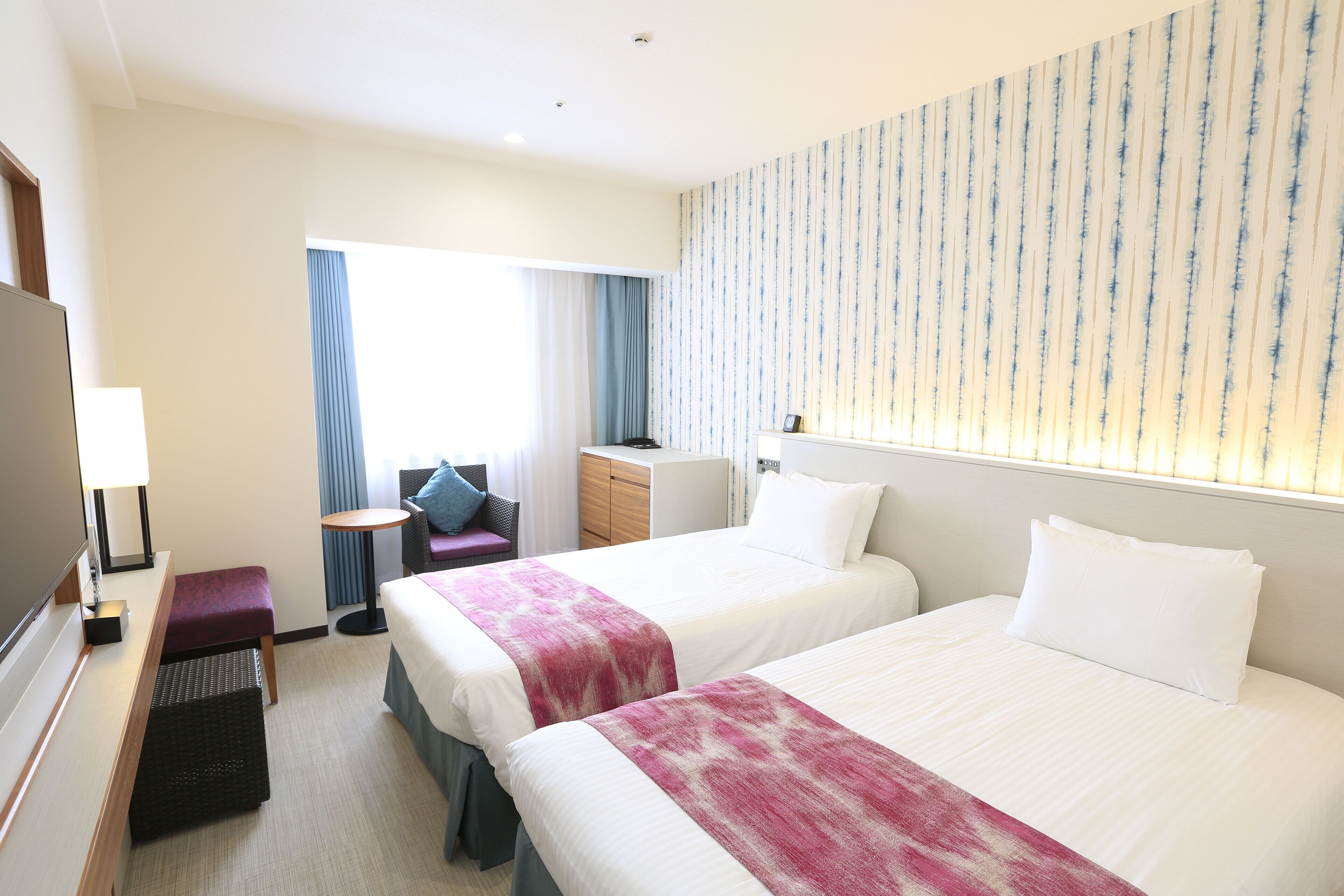 A hotel room with two beds, a desk, a chair, a small table, and a TV.