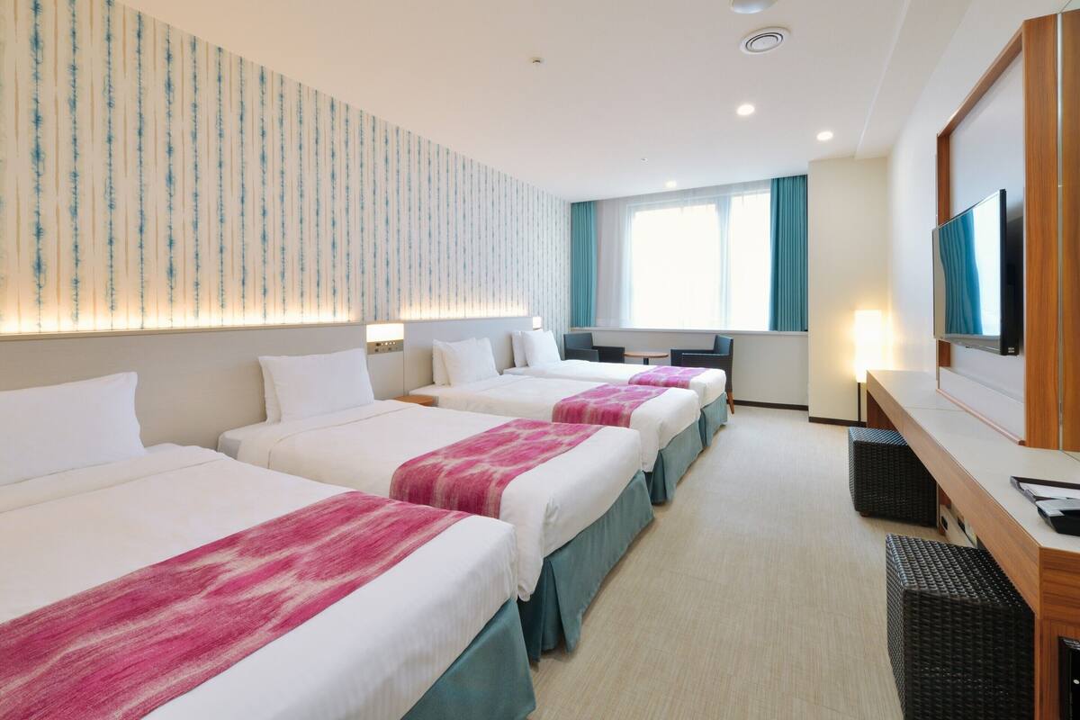 Habitación de hotel con tres camas, un escritorio, un televisor y una ventana con cortinas.