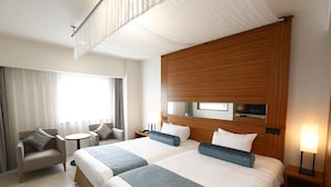 Habitación de hotel con dos camas, una zona de estar con una silla y una mesita, una mesita de noche con lámpara, y un ventanal con cortinas.