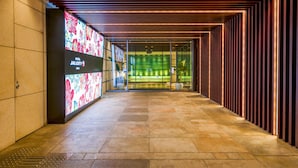 Un pasillo moderno de hotel con una pantalla digital a la izquierda, una habitación iluminada en verde al fondo y un techo con diseño.