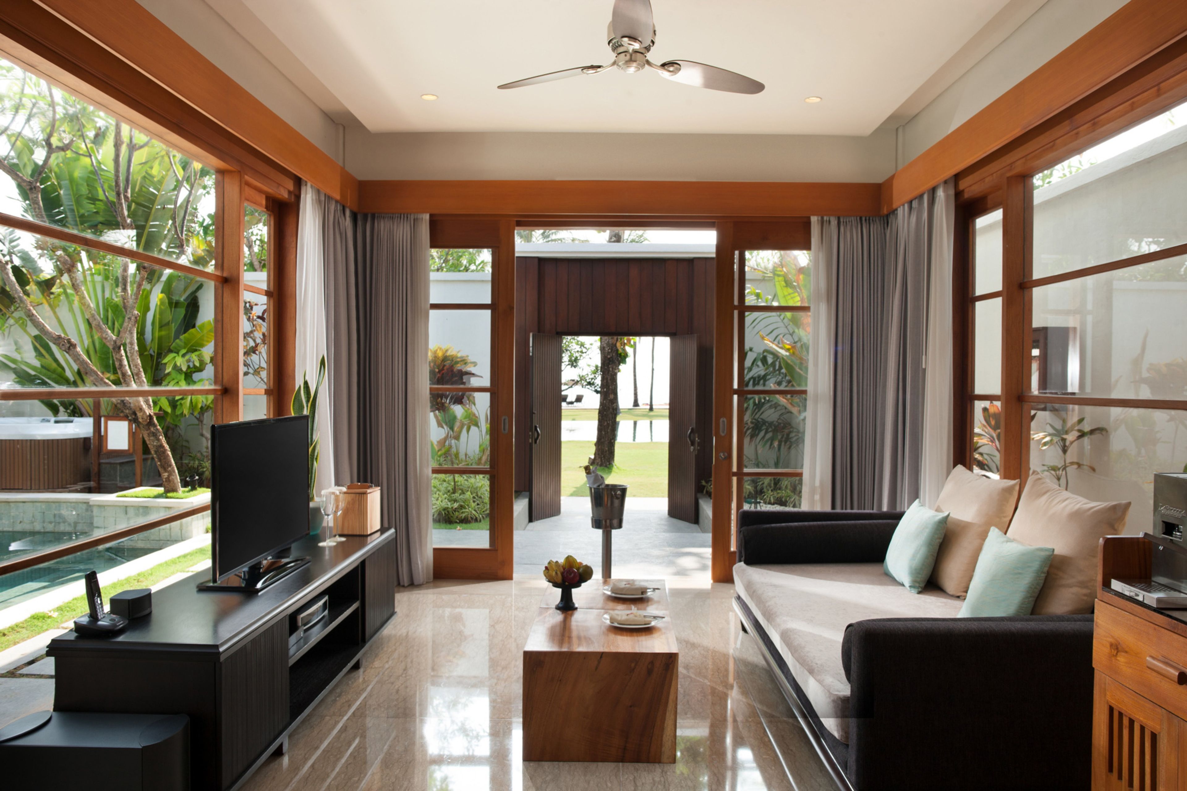Una sala de estar moderna con un televisor de pantalla plana, una mesa de centro de madera y un sofá con cojines. Hay un separador de vidrio que ofrece vistas a una piscina y follaje exterior.