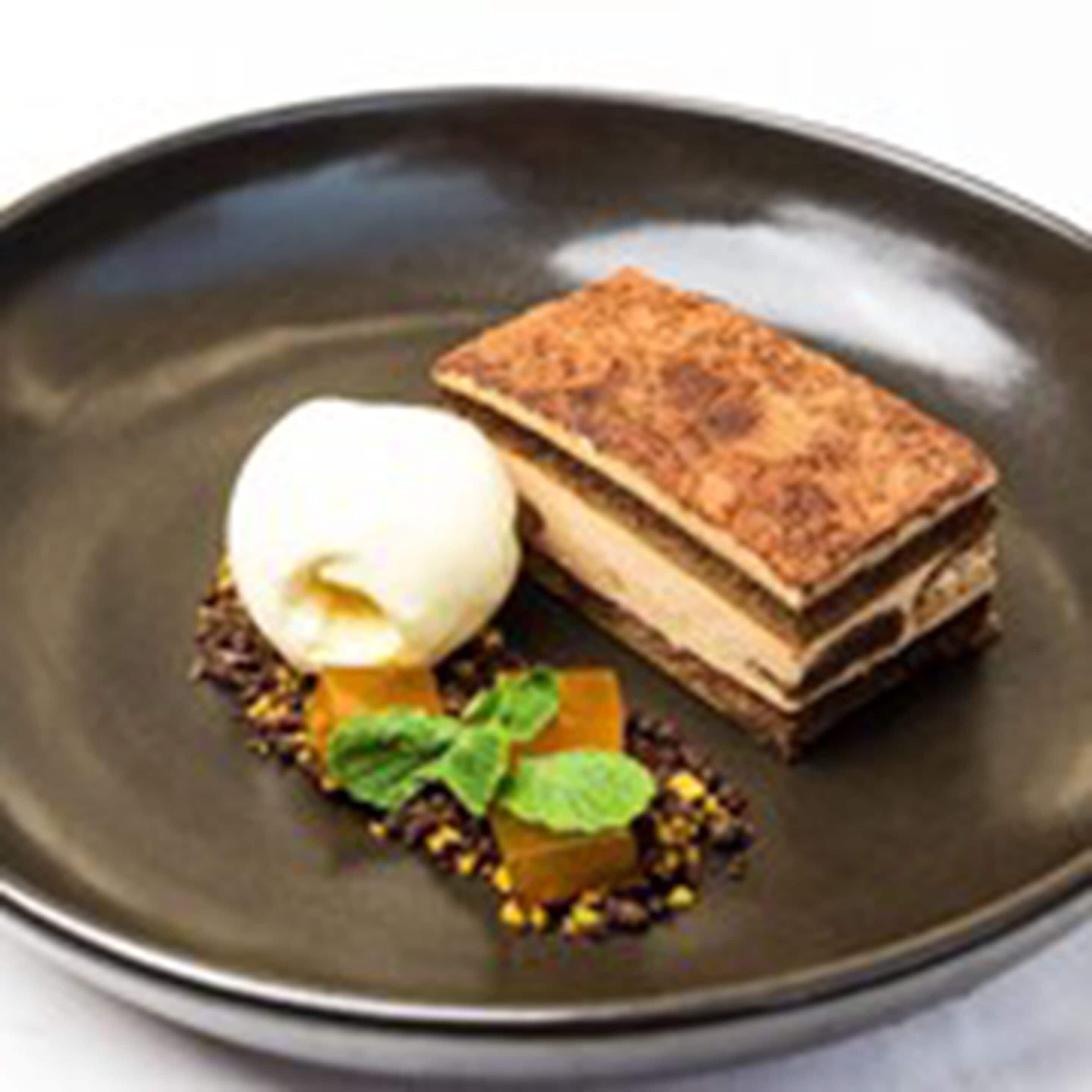 Un postre con una tarta en capas y una bola de helado sobre un plato oscuro.