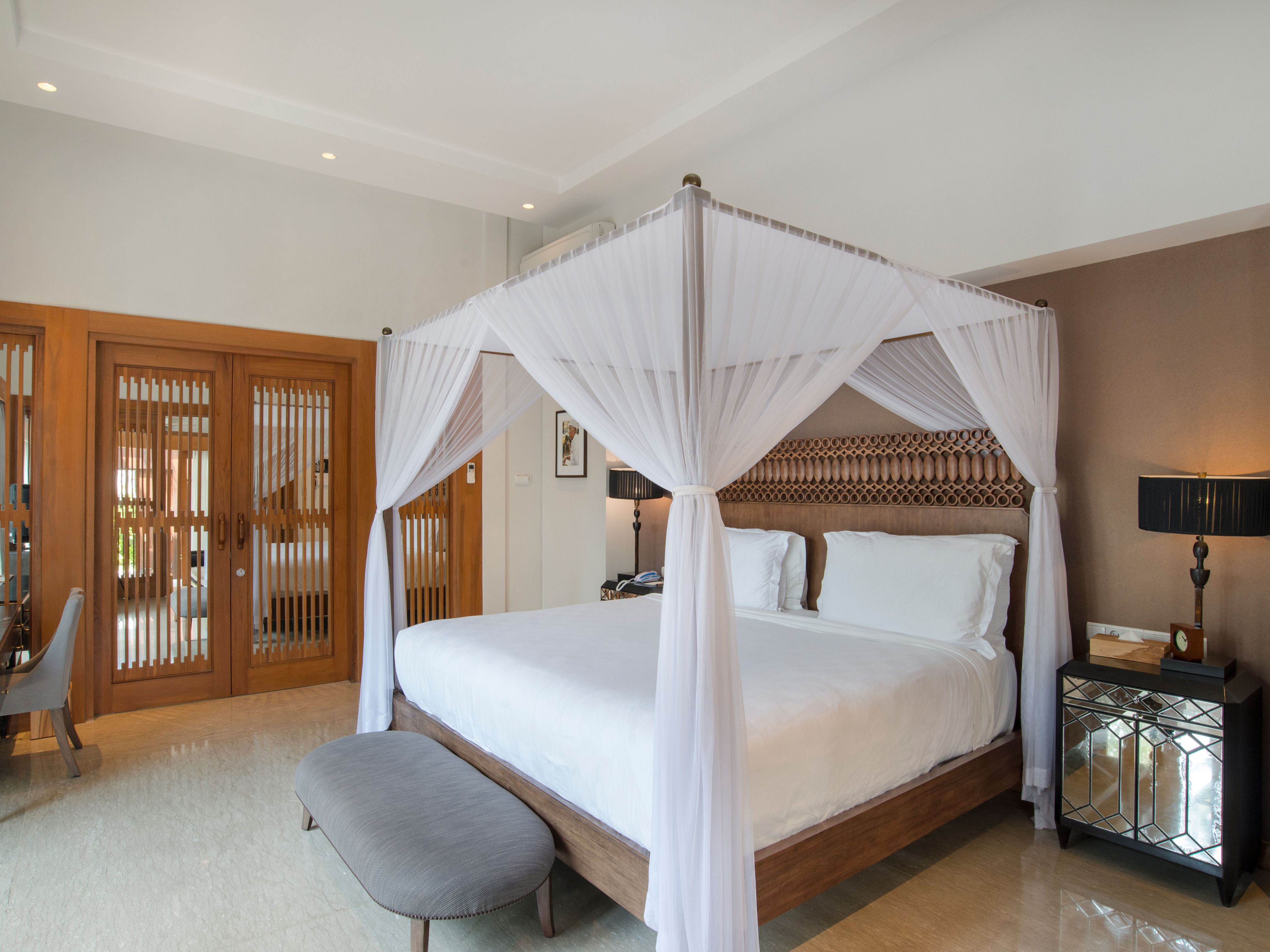 A four-poster canopy bed with a wooden headboard, a bedside table with a clock, and a bench at the foot of the bed.