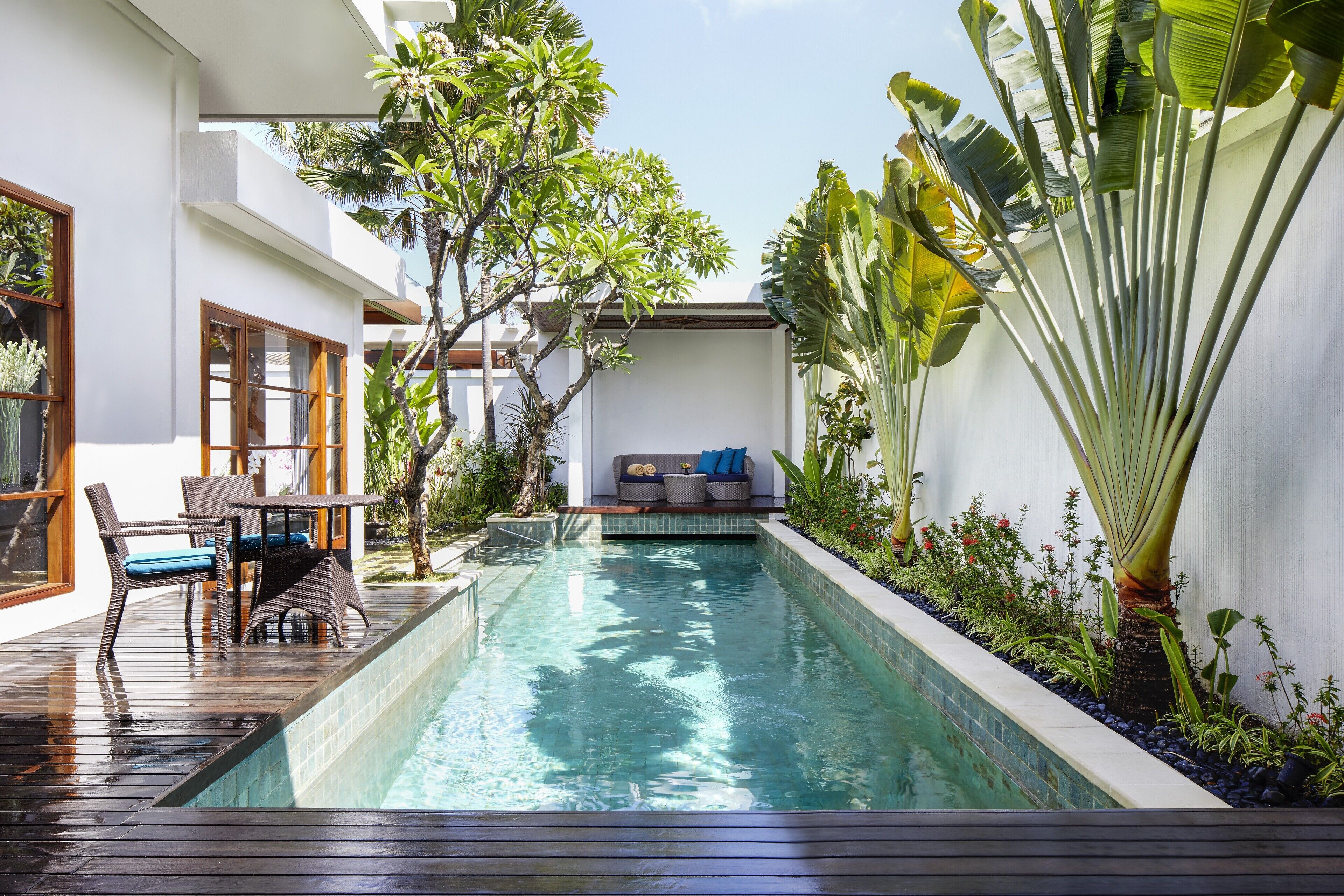 A modern outdoor pool area with a wooden deck, seating, and lush tropical plants.