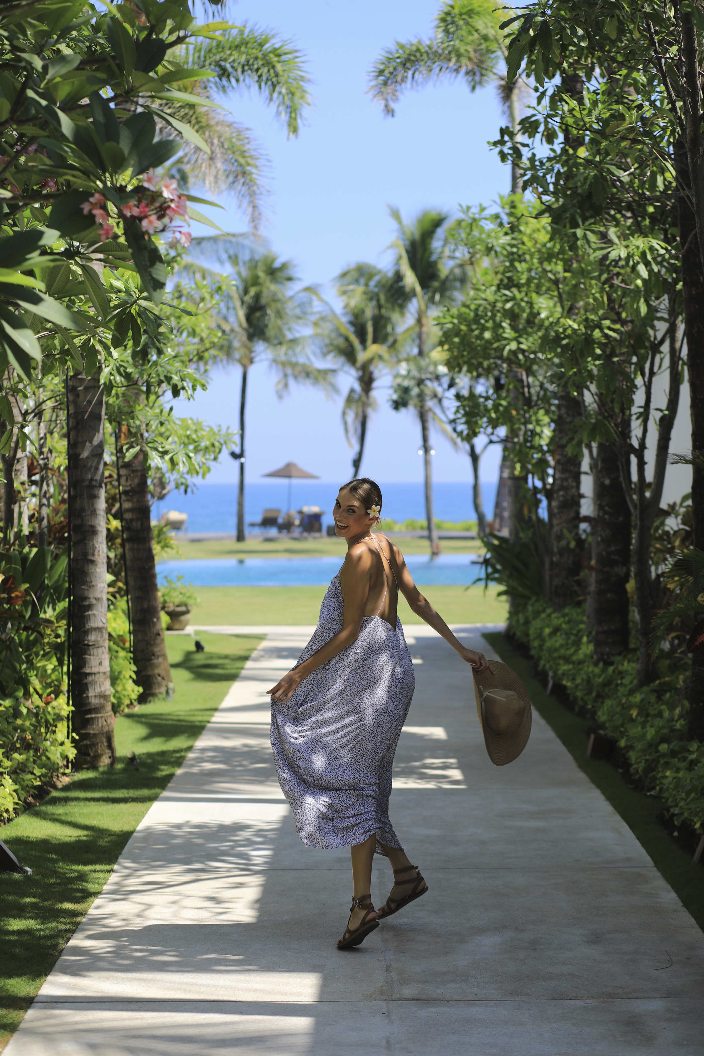 A pathway lined with palm trees and greenery, leading to a pool area with a view of the ocean.