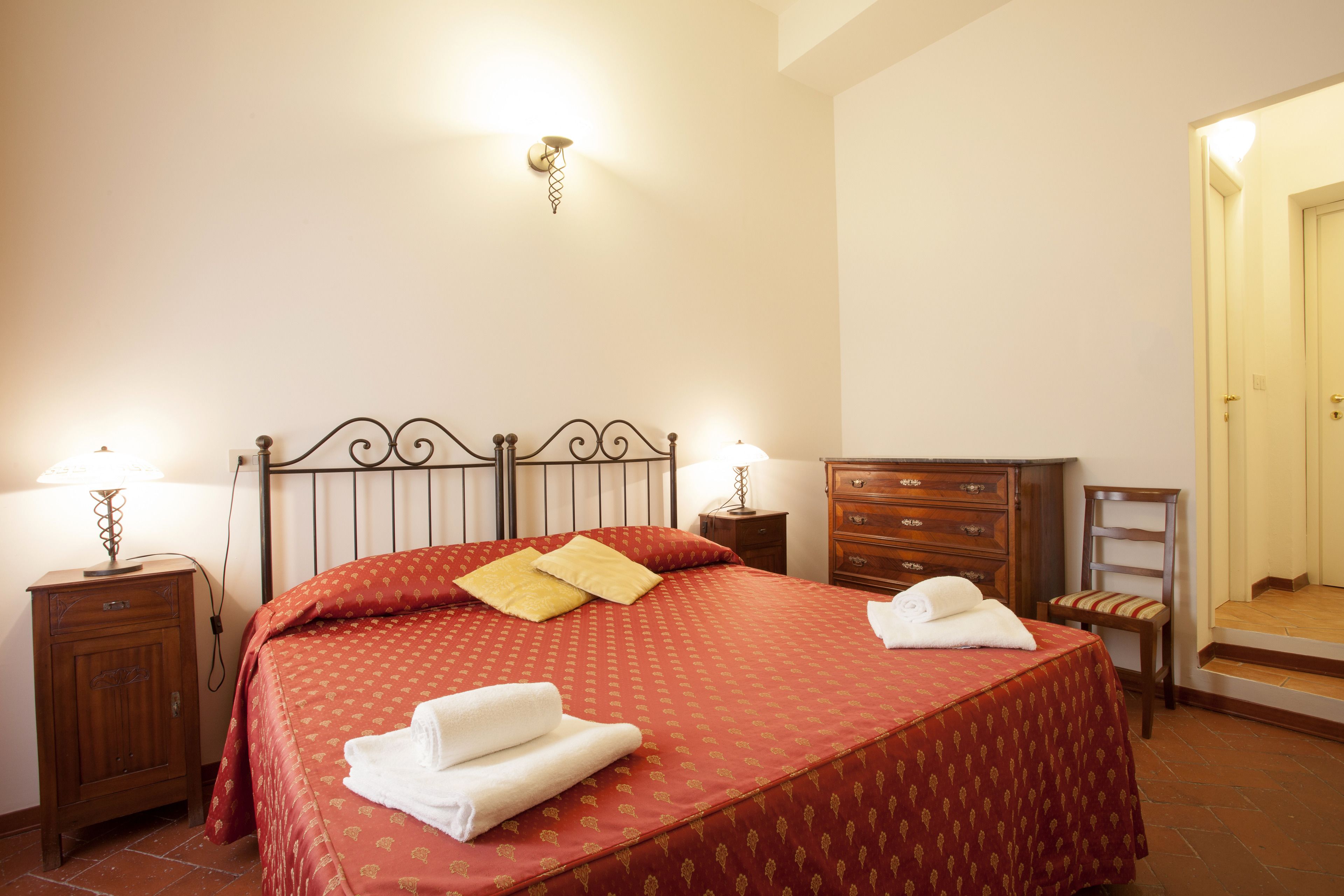 Una cama bien hecha con una colcha roja, dos almohadas y dos toallas dobladas. Hay dos mesitas de noche con lámparas, un aparador de madera y una silla.