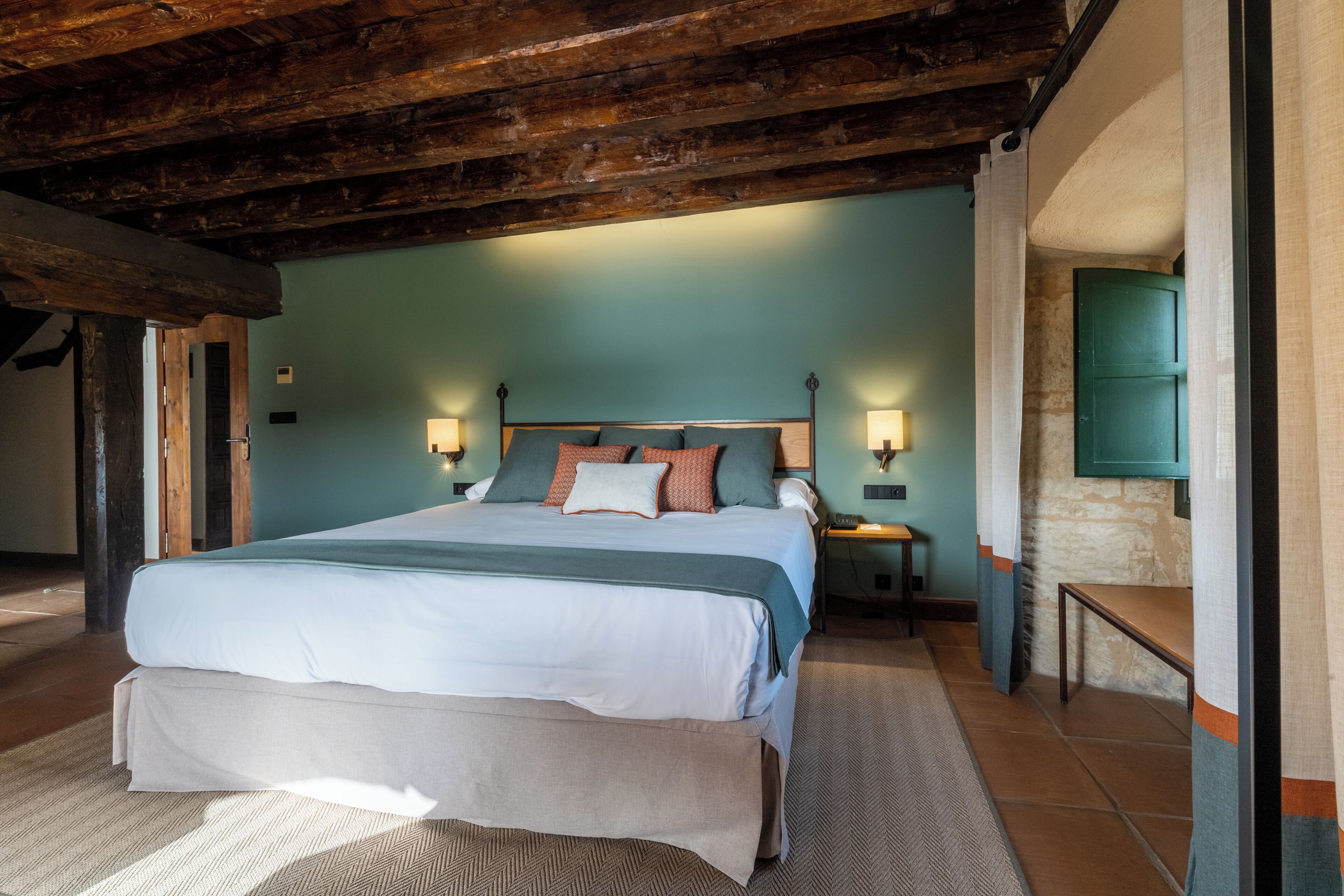 Une chambre avec un grand lit, des poutres en bois, un mur en pierre et une porte verte.