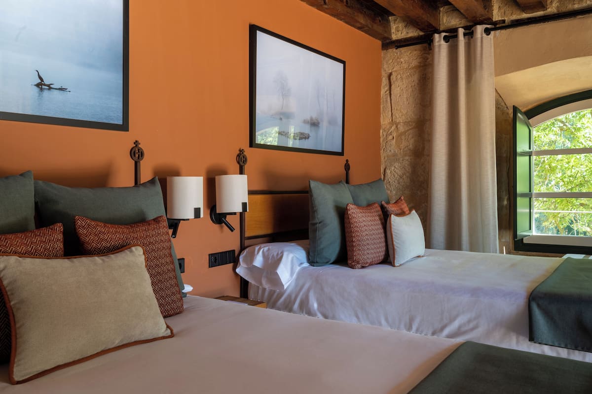 Habitación de hotel con dos camas, una mesita de noche, una lámpara, un cuadro enmarcado y una ventana con vistas.