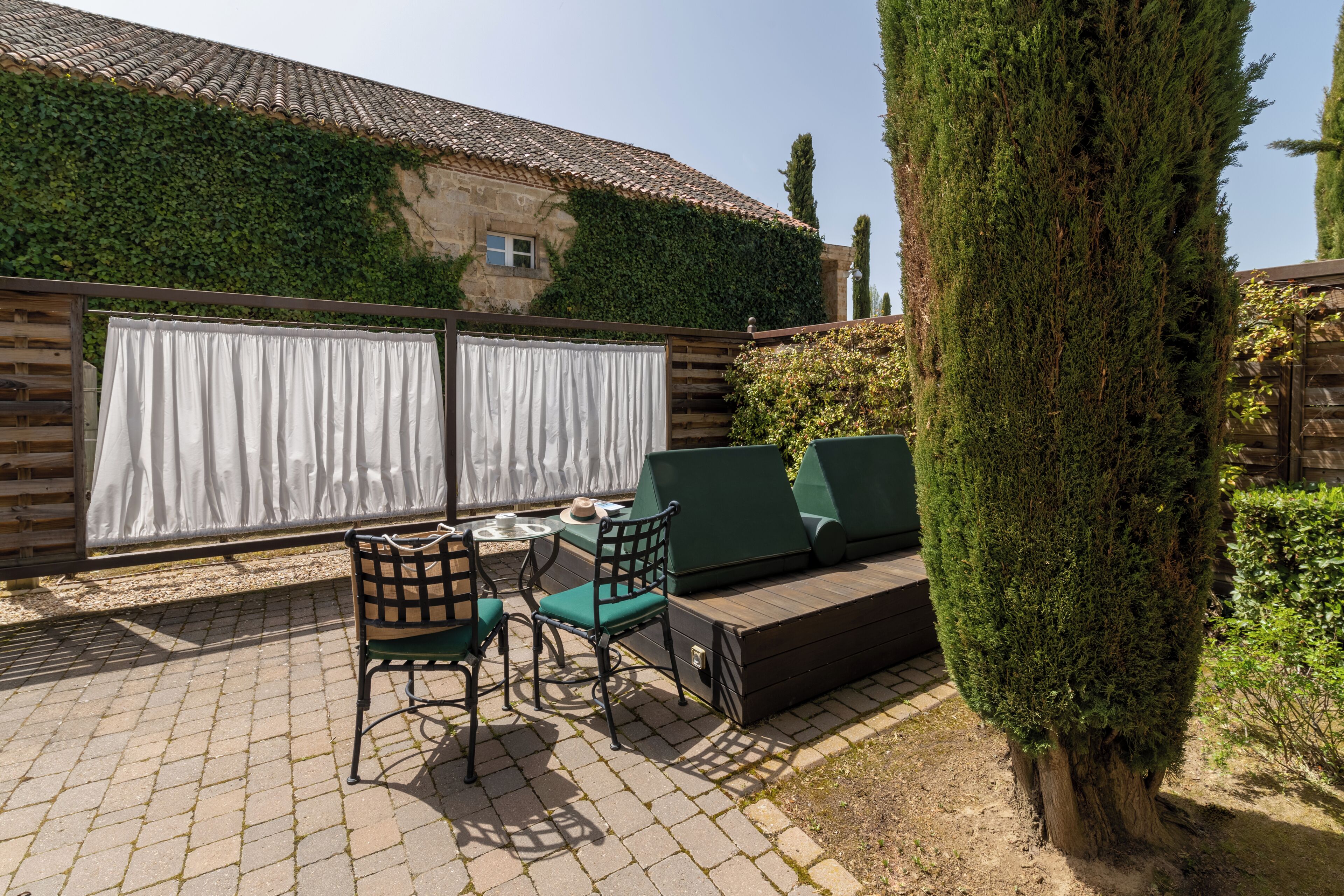 Eine Terrasse mit Tisch und Stühlen, ein grünes Sofa und ein hoher Baum.