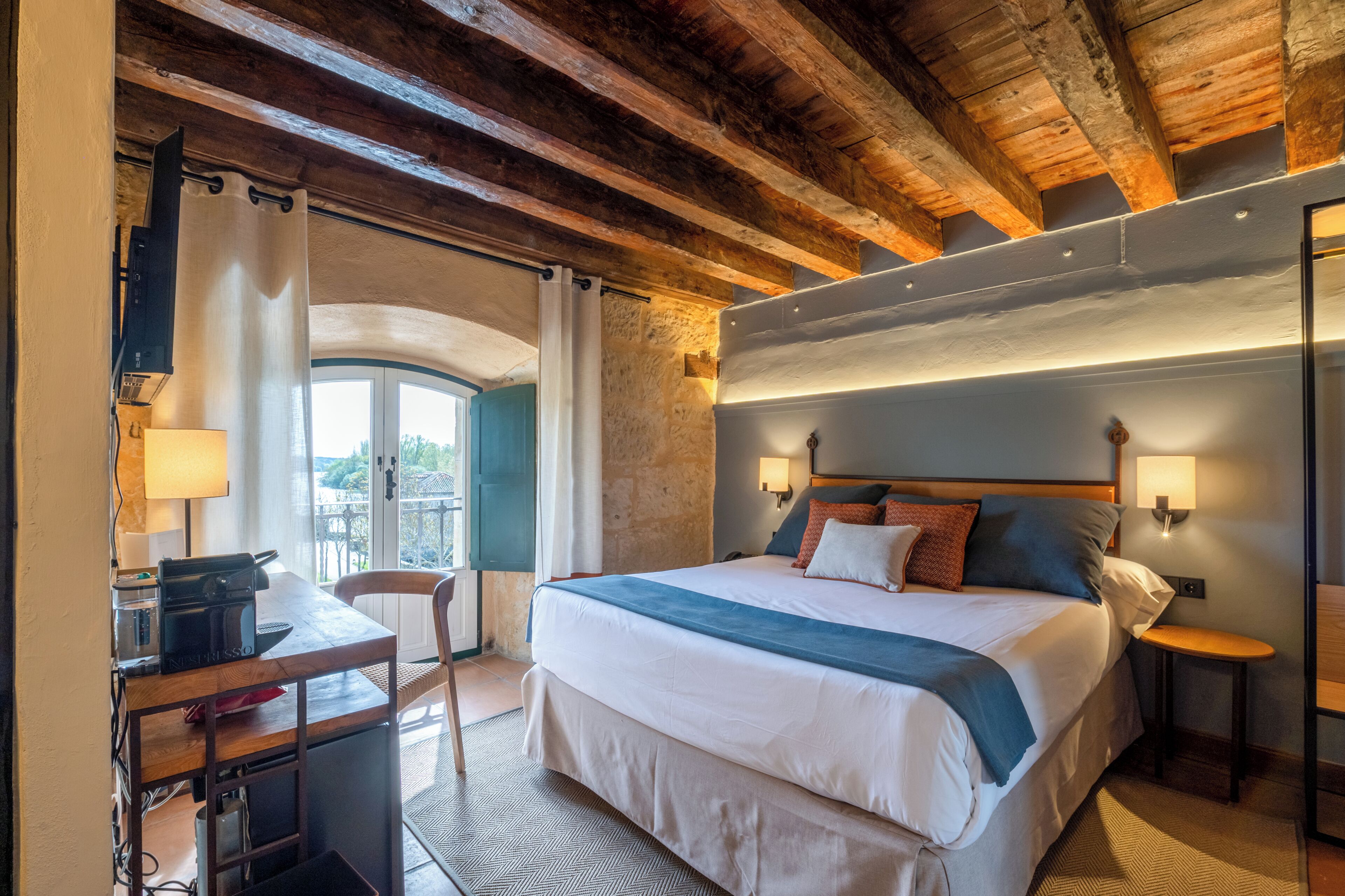 Een hotelkamer met een bed, nachtkastjes, een bureau met een koffieapparaat, een stoel en uitzicht op het landschap.
