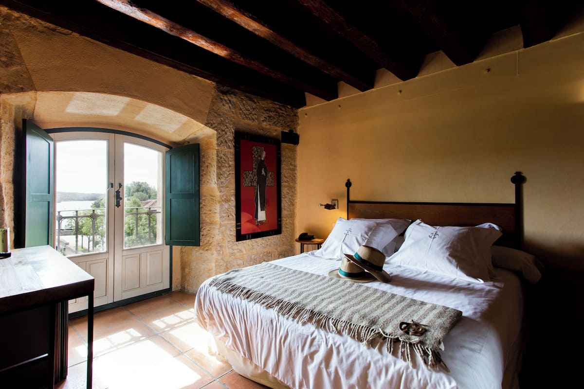 Un dormitorio con cama, ventana, mesa y vista al exterior.