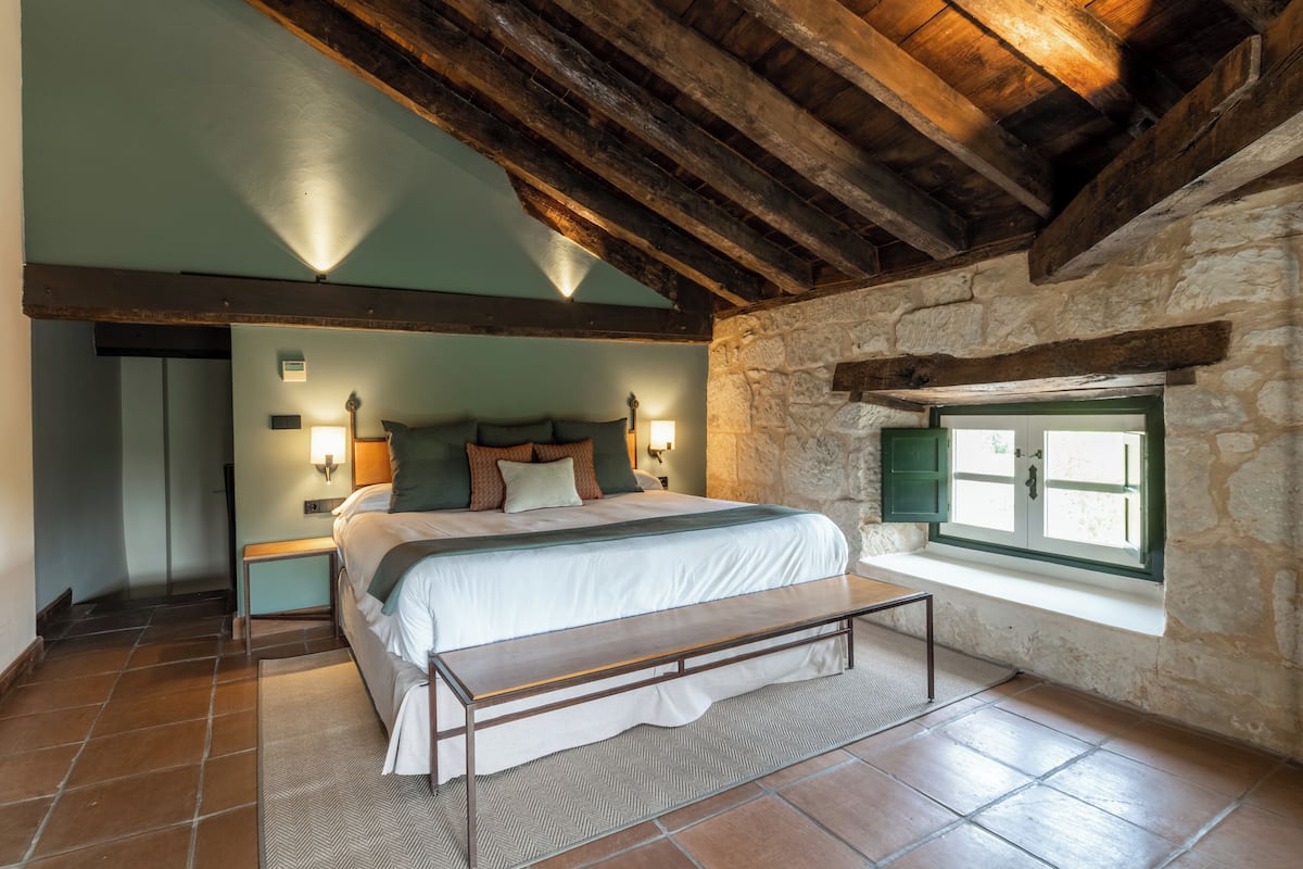 Un dormitorio con cama, mesitas de noche, un banco, una pared de piedra y una ventana.