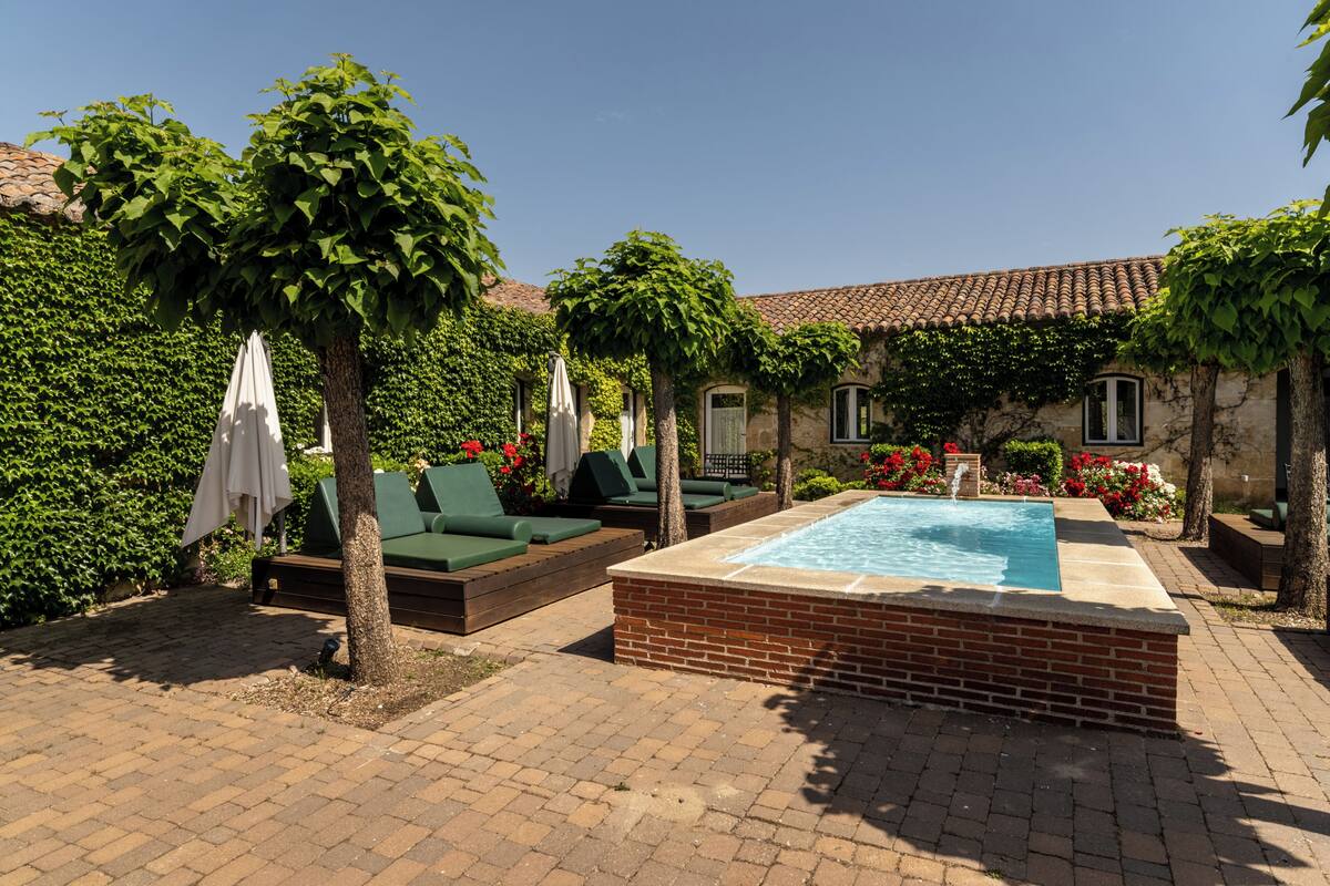 Un patio con piscina, sillones de descanso verdes y un edificio de piedra cubierto de hiedra.