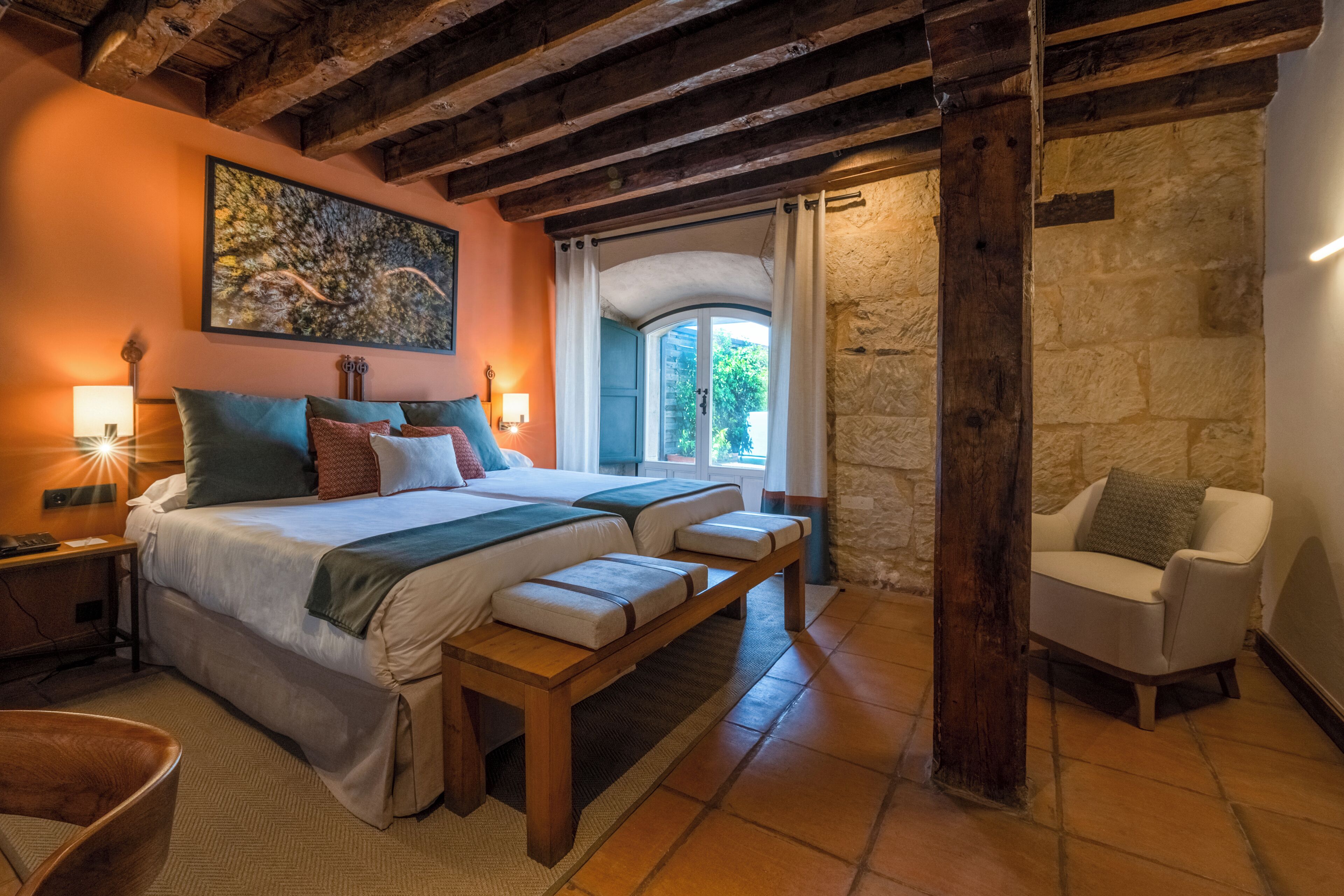 Un dormitorio con una cama grande, mesitas de noche, un banco, una silla, un muro de piedra y una ventana con cortinas.