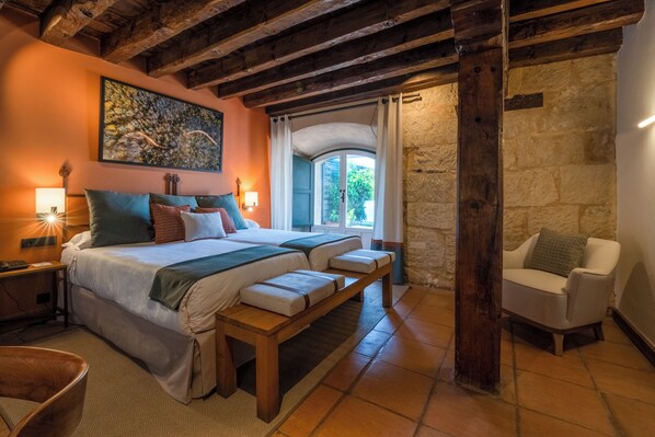 Un dormitorio con una cama grande, mesitas de noche, un banco, una silla, un muro de piedra y una ventana con cortinas.