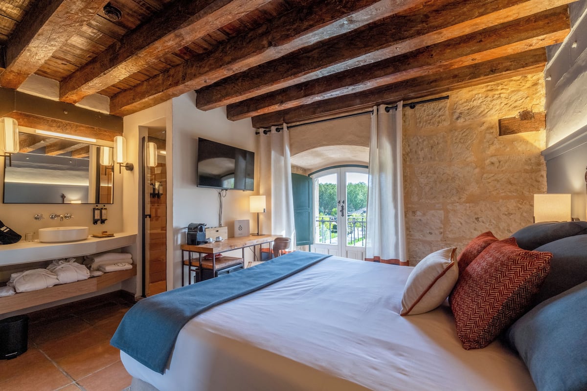 Habitación de hotel con techo de madera, cama con almohadas, escritorio con silla, baño con lavamanos y espejo, y vistas a zonas verdes.
