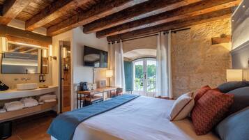 Habitación de hotel con techo de madera, cama con almohadas, escritorio con silla, baño con lavamanos y espejo, y vistas a zonas verdes.