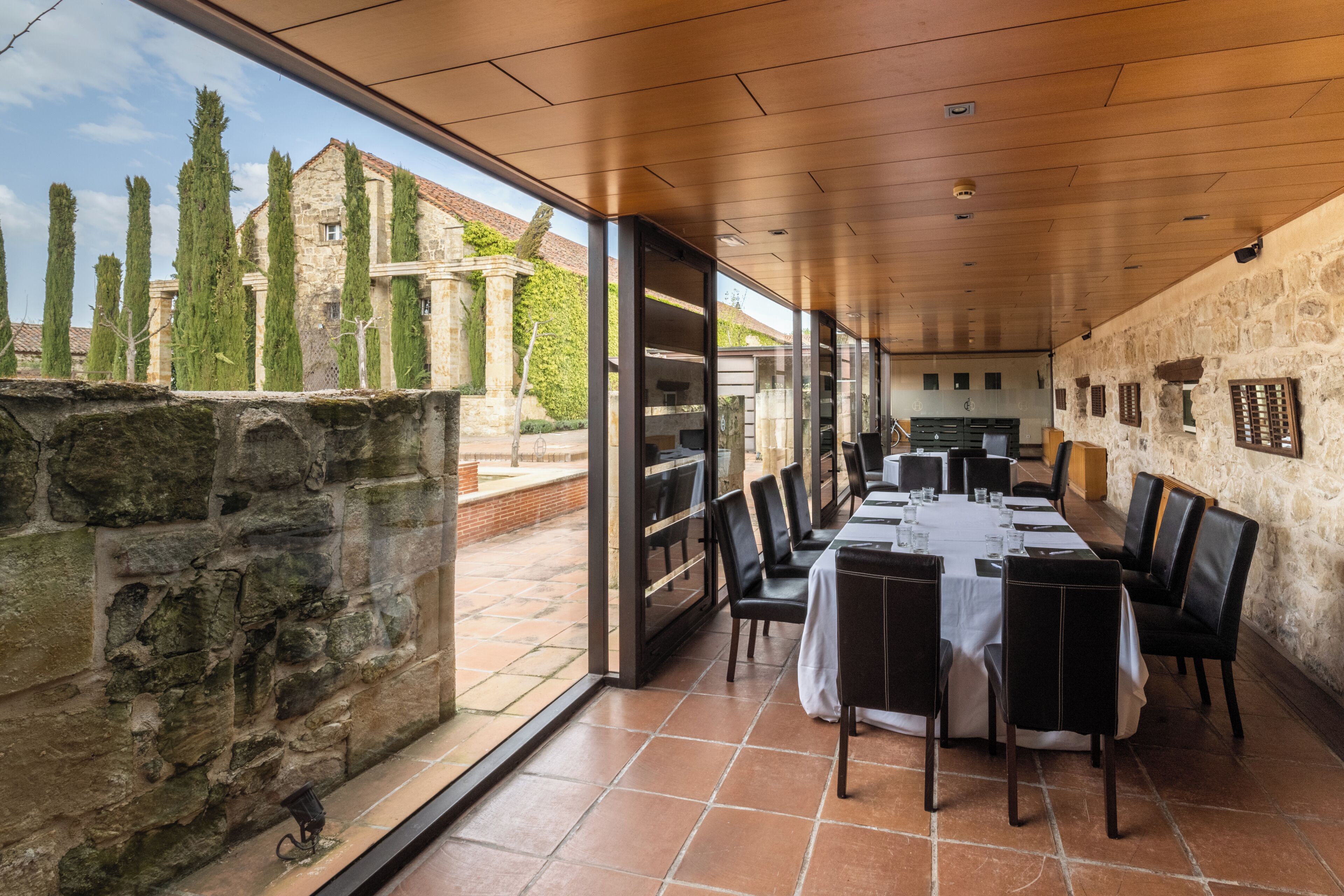 Un restaurante con una larga mesa, sillas, paredes de piedra y vistas a árboles y edificios.