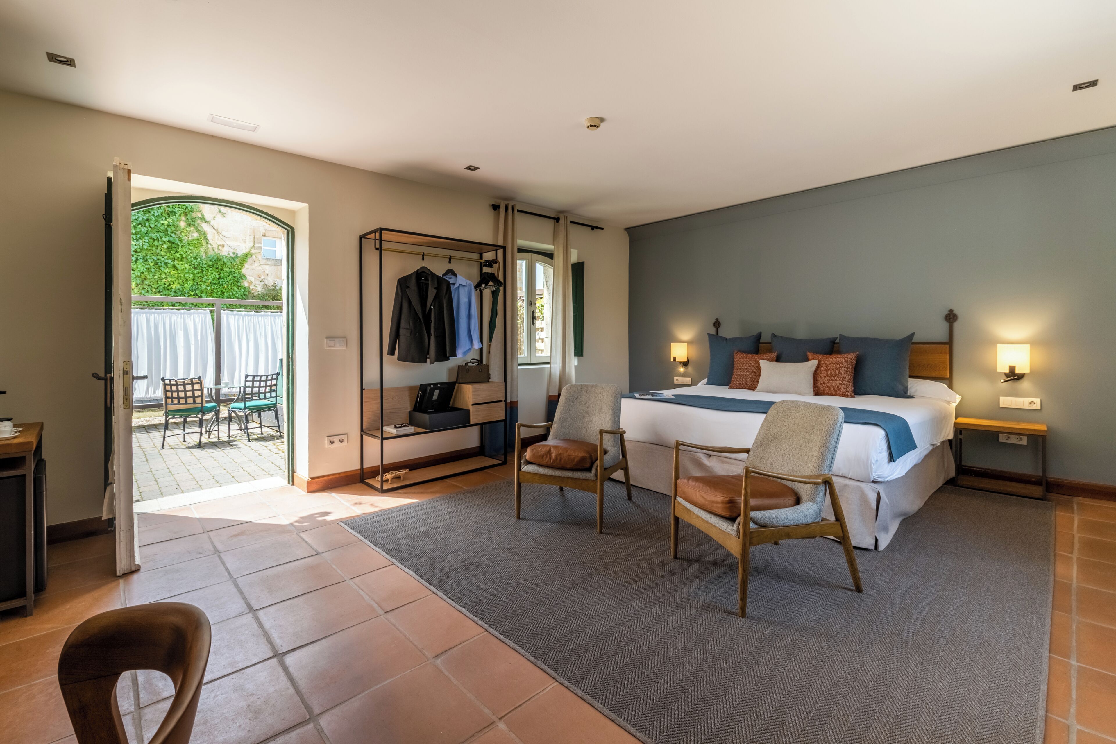 A bedroom with a bed, two chairs, a desk, a wardrobe, and a view of the outside.