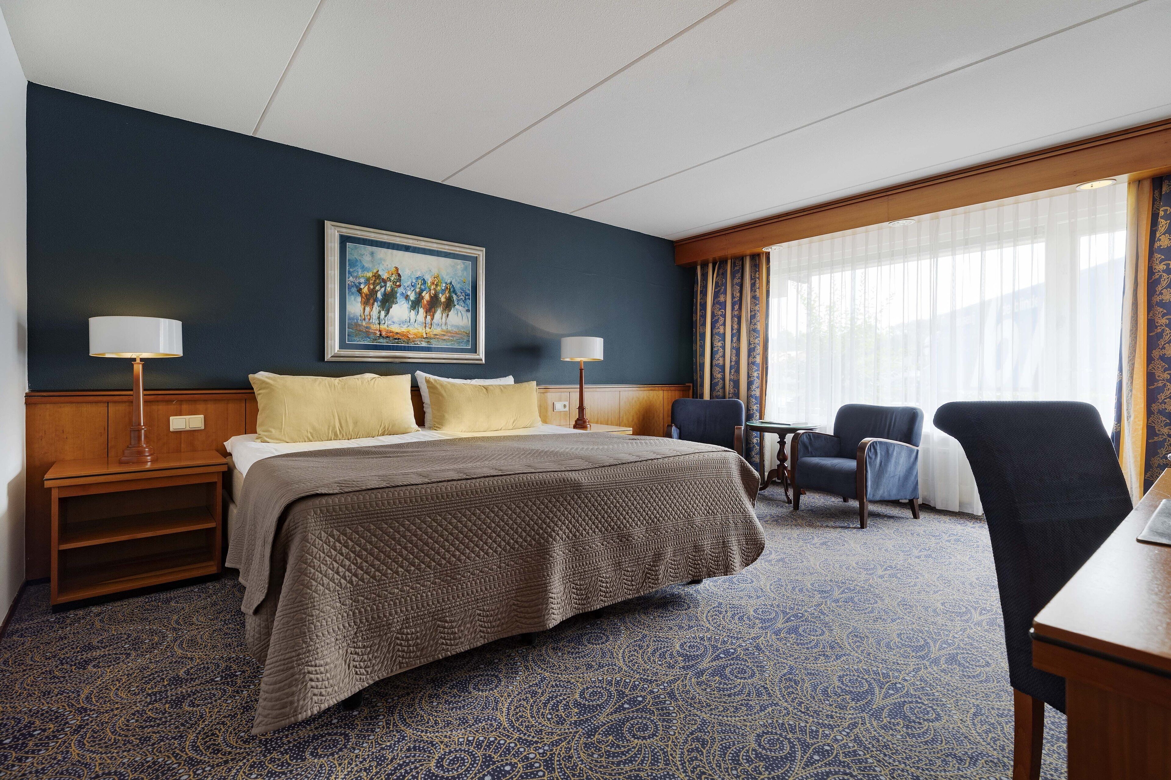 Habitación de hotel con una cama grande, dos mesitas de noche, un escritorio, una silla y una ventana con cortinas.