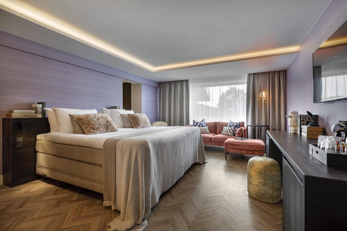 Une chambre d’hôtel moderne avec un grand lit, un coin salon doté d’un canapé rose, une petite table avec une lampe et un téléviseur fixé au mur.