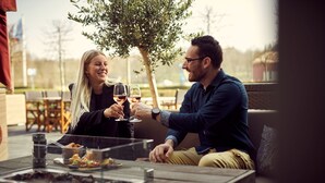 Ein Paar, das mit Weingläsern anlässt, sitzt an einem Tisch im Freien, neben einem Glastischschrank, umgeben von einer ungezwungenen Gastronomie-Einrichtung.