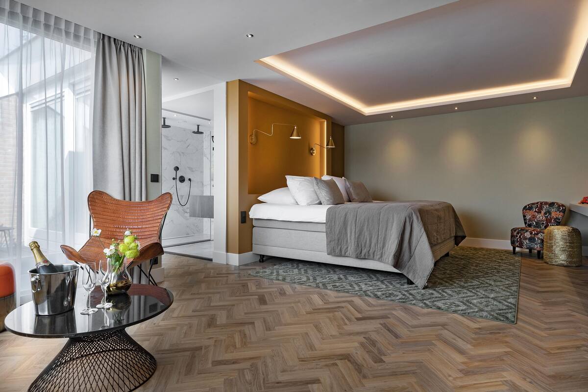 Une chambre d’hôtel moderne avec un lit, une table à manger avec une bouteille et des verres, une chaise et un espace douche.