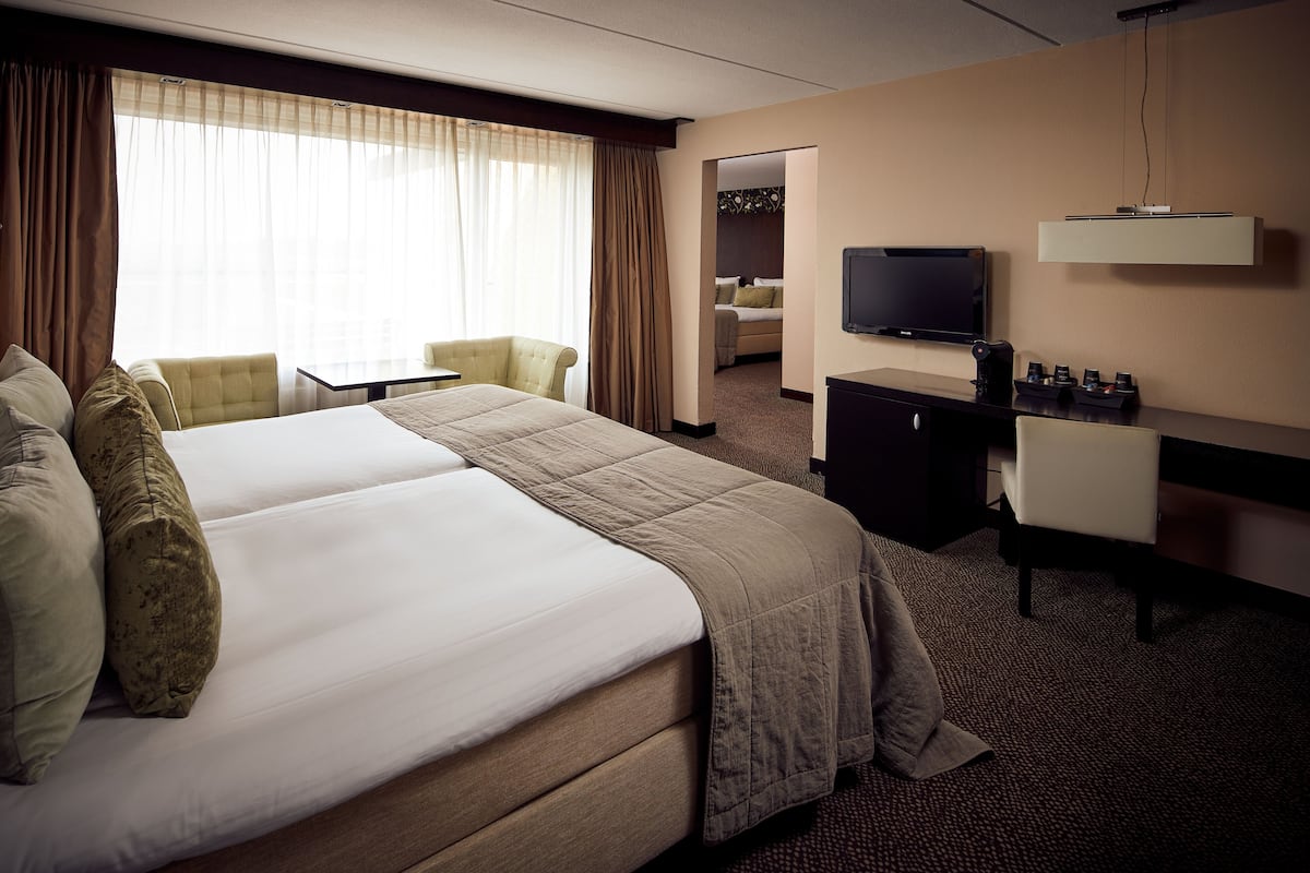 Une chambre d’hôtel avec un grand lit, un bureau avec un ordinateur, une télévision et un coin salon avec des fauteuils.