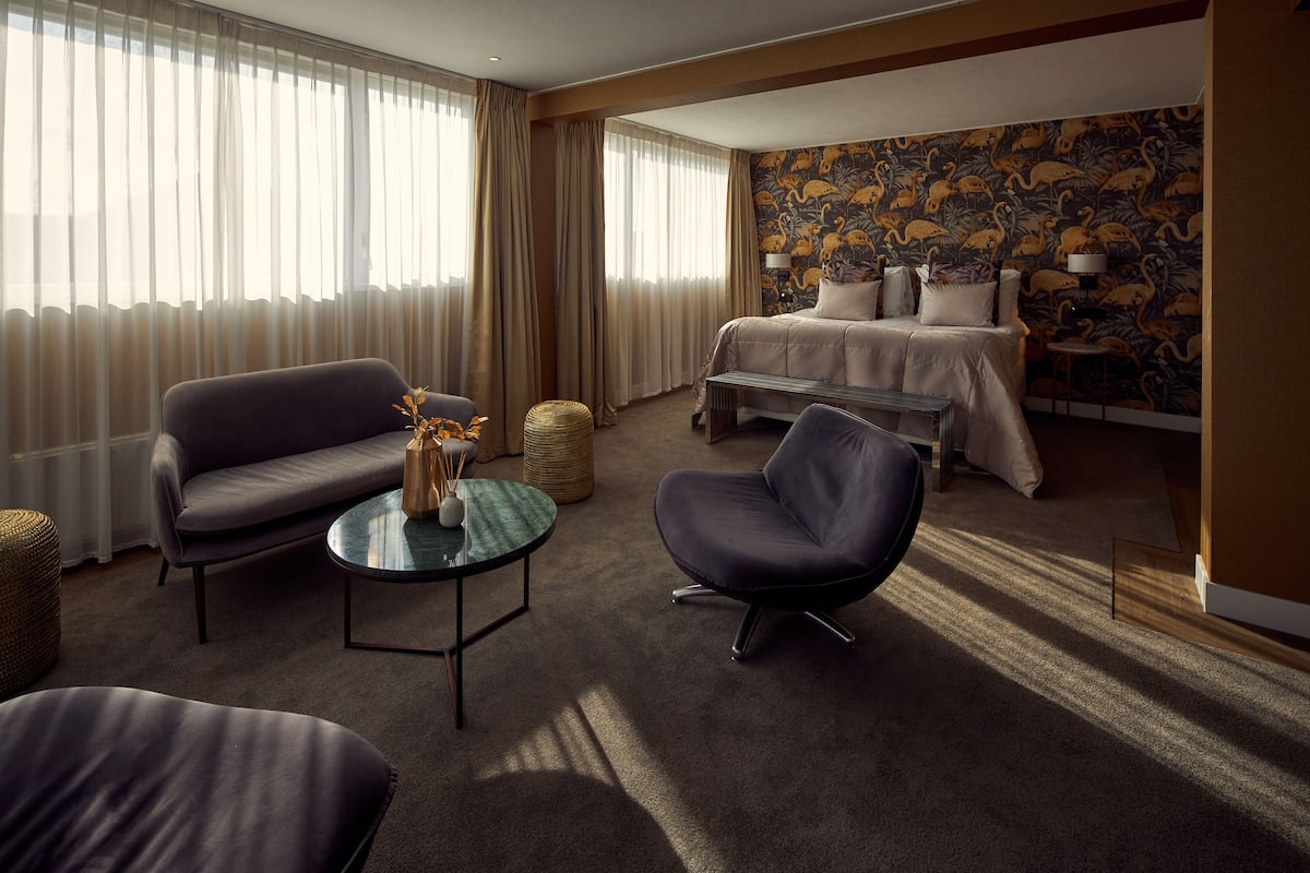 Une chambre d’hôtel avec un canapé, un fauteuil et un lit dont le mur est recouvert d’un papier peint à motifs floraux.
