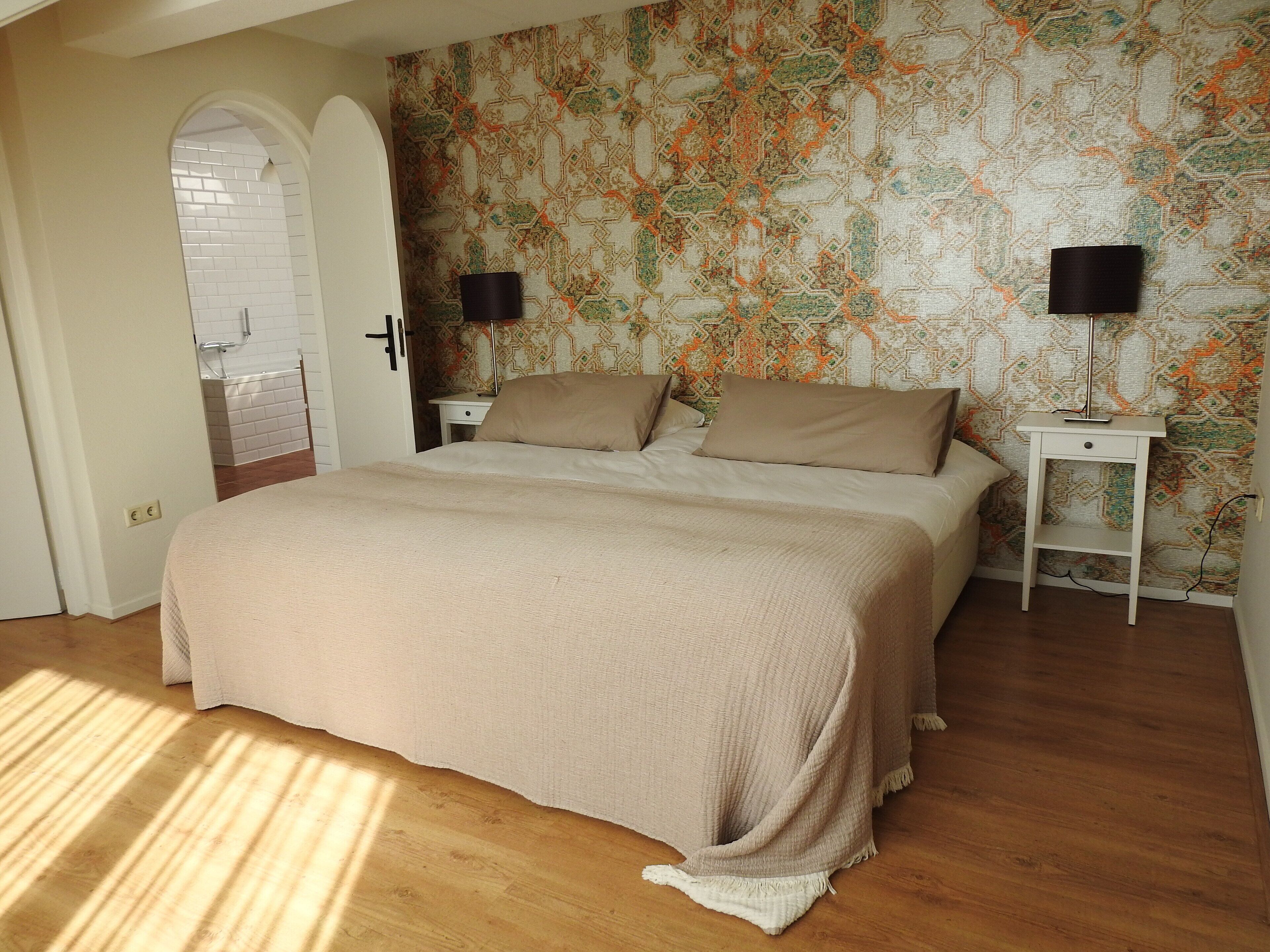 Un dormitorio con una cama grande, dos mesitas de noche con lámparas, un papel tapiz estampado y suelo de madera.