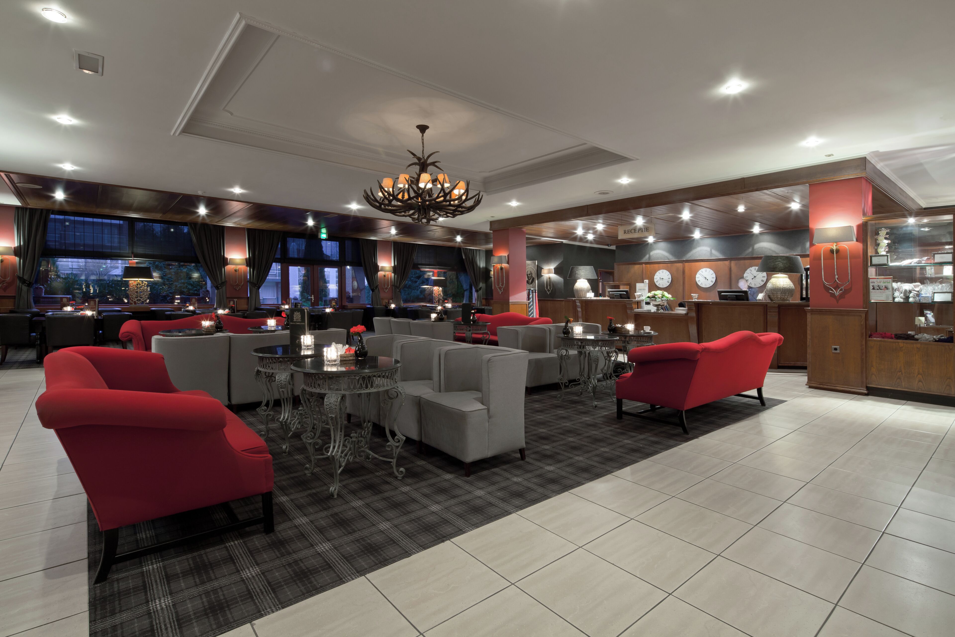 A modern hotel lobby with red and grey seating, a central chandelier, and a reception desk.