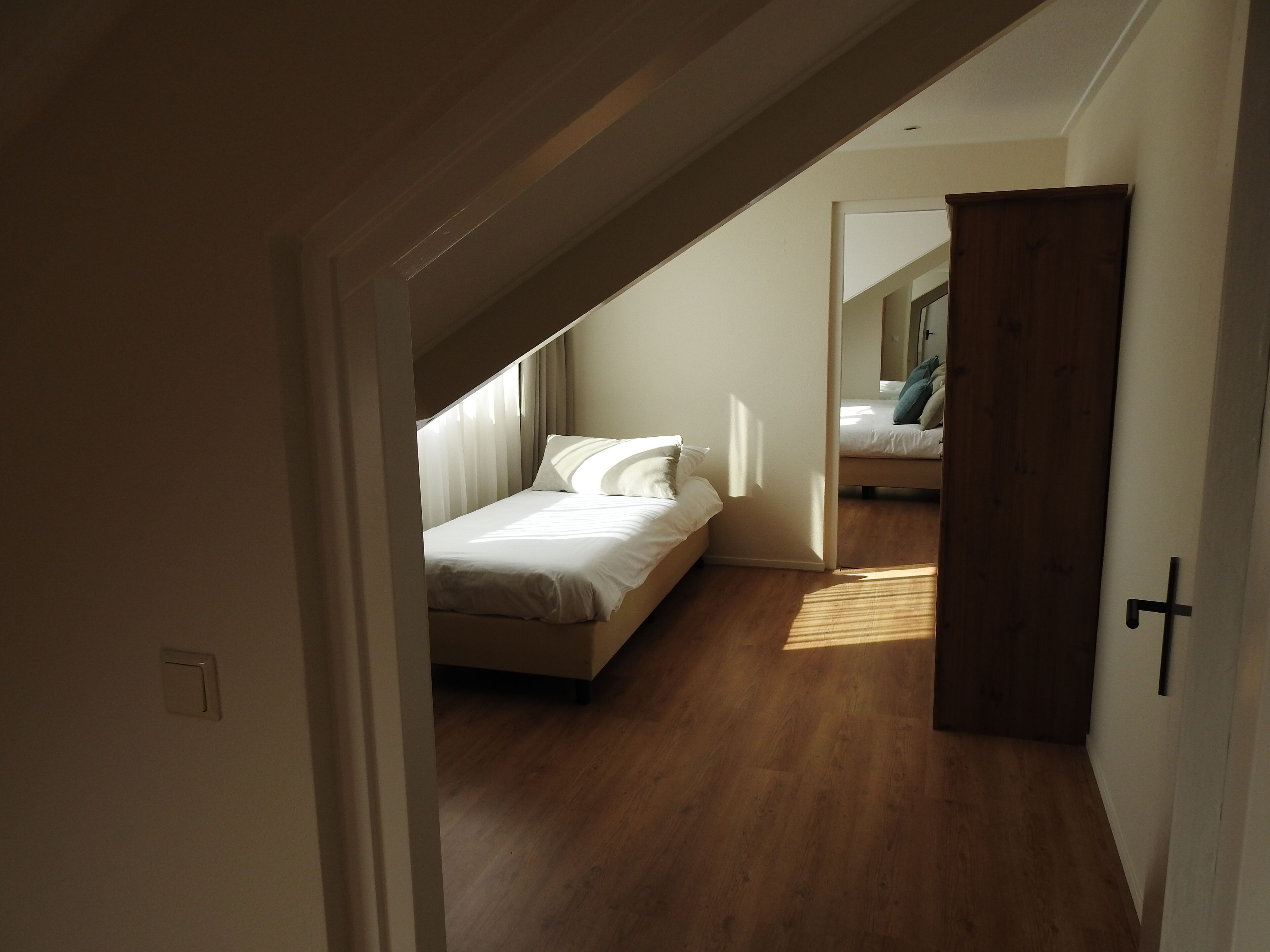 A bedroom with a bed, a wooden floor, and a door leading to another room.