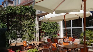 Een terras buiten met tafels en stoelen, omringd door plantenpotten en een witte parasol.