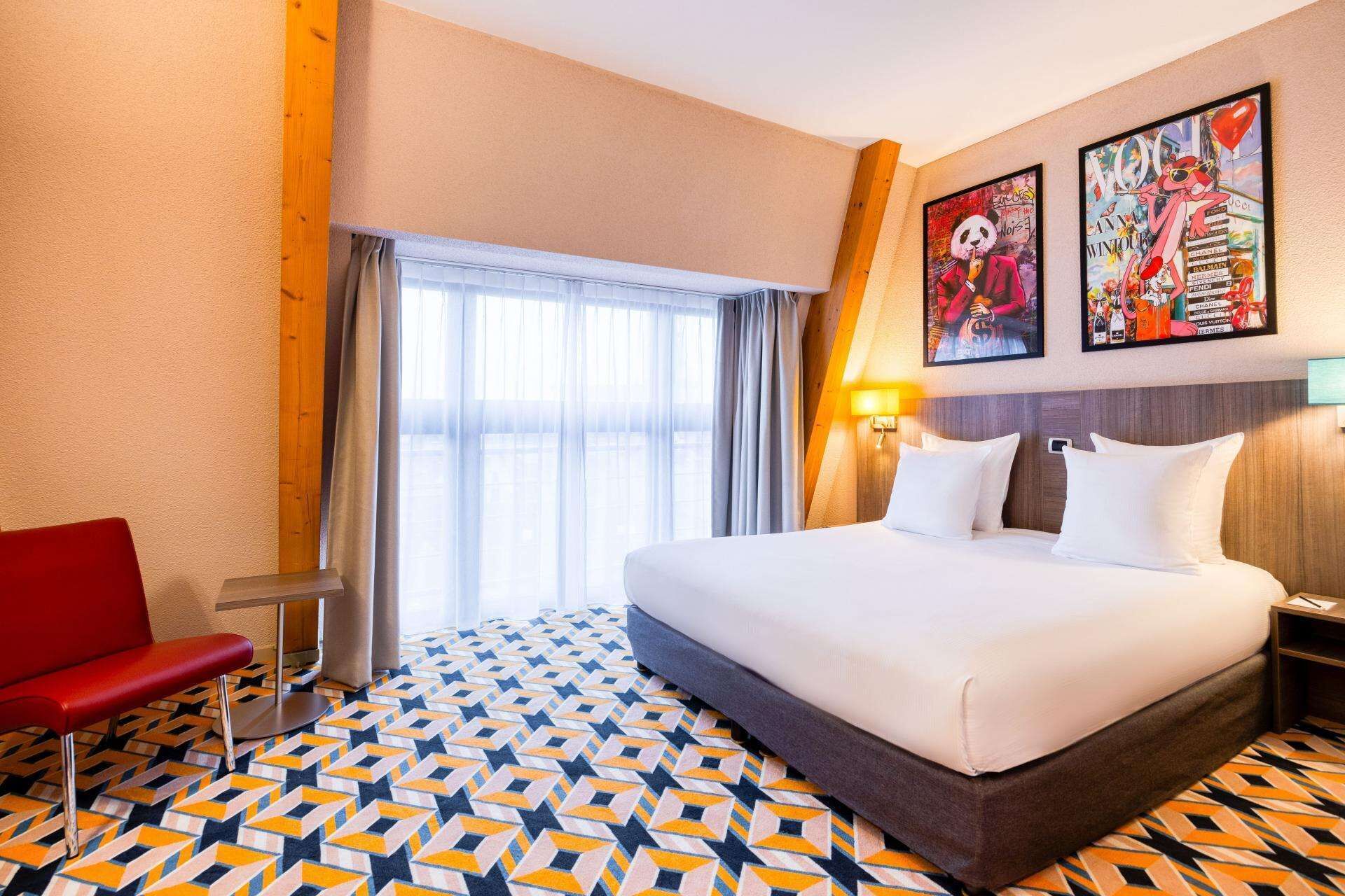 A hotel room with a large bed, a red chair, and two wall art pieces.