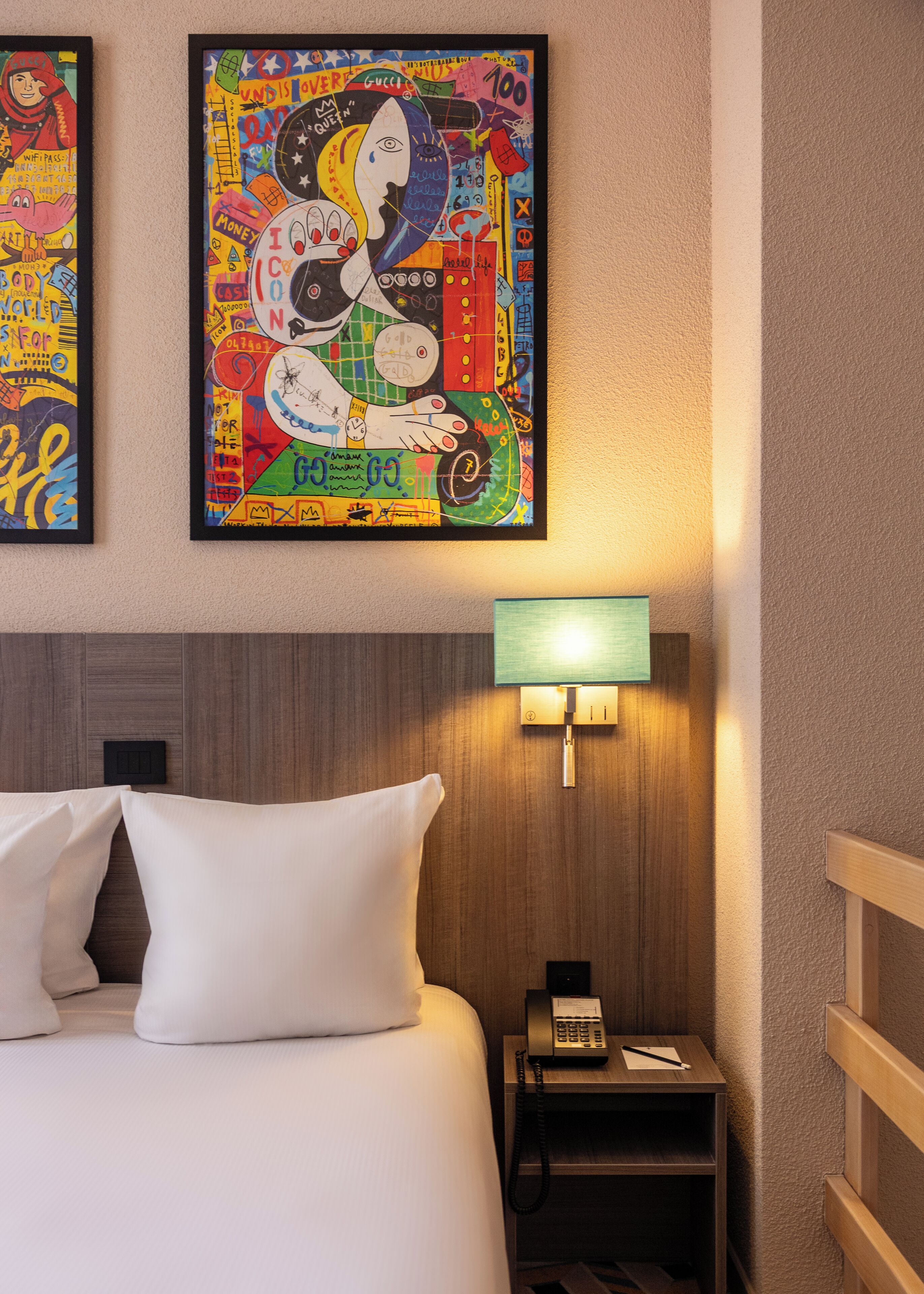 A hotel room with a bed, a nightstand with a lamp, and two colorful artworks on the wall.