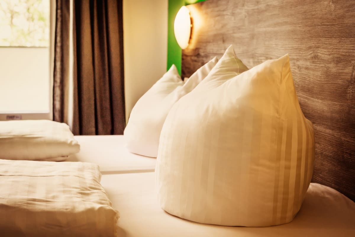 A neatly made bed with white bedding and pillows, a wooden headboard, and a window with curtains.