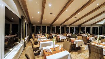 Een restaurant met meerdere tafels en stoelen, grote ramen en een hoge plafond met zichtbare balken.