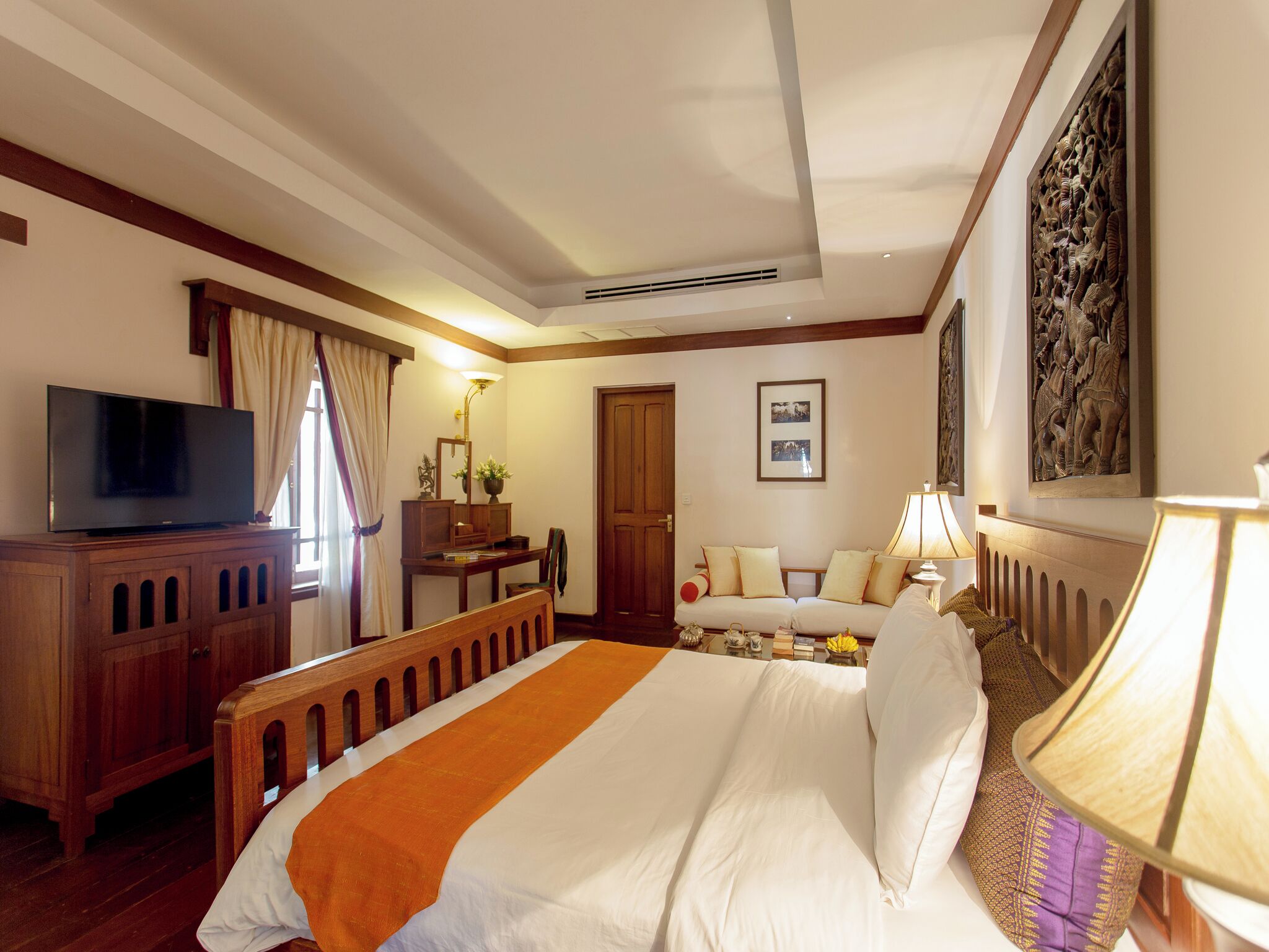 A hotel room with a bed, a desk with a TV, a sofa, a lamp, and artwork on the walls.