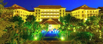 Un hotel grande de varios pisos con una fachada simétrica, una entrada central y un jardín bien iluminado con palmeras.