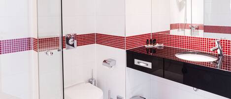 Un baño moderno con cabina de ducha de vidrio, inodoro blanco y lavamanos negro con salpicadero de azulejos rojos.