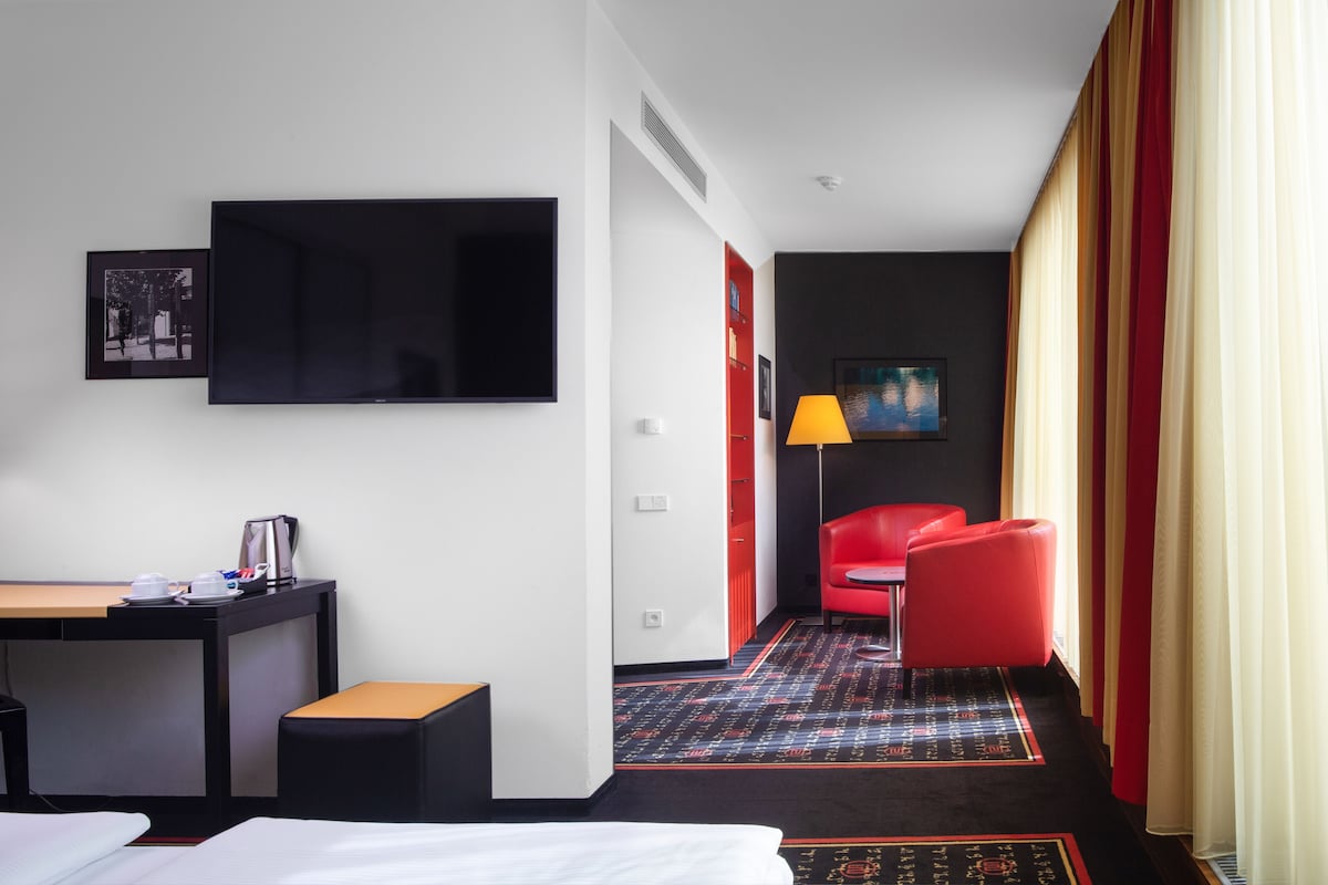 Une chambre d’hôtel avec un lit, un bureau doté d’une machine à café, une télévision, un fauteuil rouge et un tableau accroché au mur.