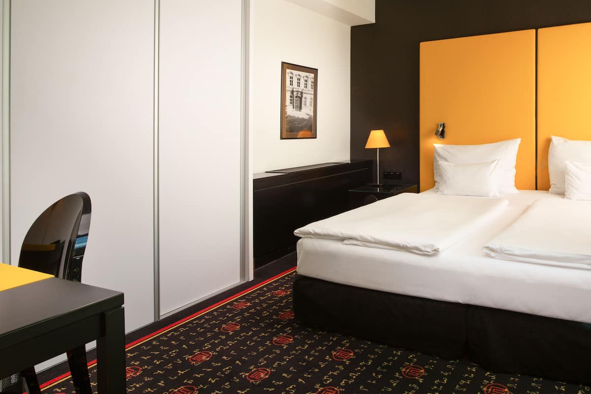 Une chambre d’hôtel avec un lit soigneusement fait, un bureau avec une chaise, une lampe et un tableau encadré au mur.