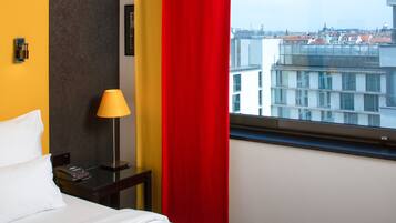 Habitació d'hotel amb un llit, una meseta amb una làmpada i una finestra que mostra una vista urbana.