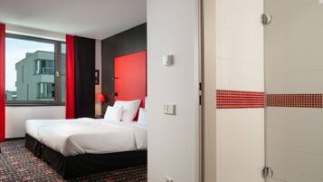 A hotel room with a bed, a red accent wall, a window with curtains, and a tiled shower area.