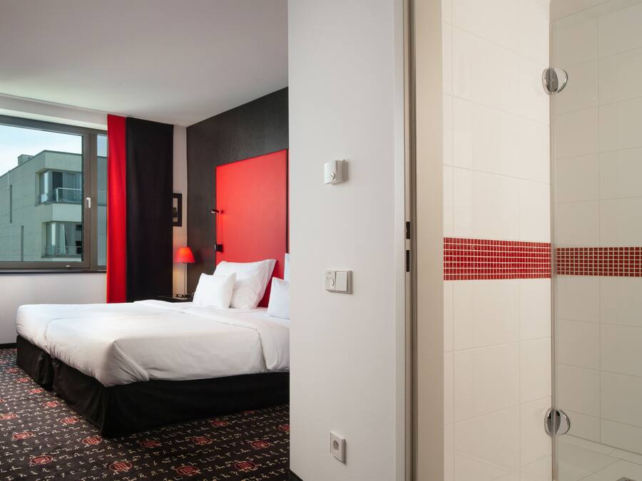 Habitación de hotel con cama, pared de acento roja, ventana con cortinas y zona de ducha con azulejos.